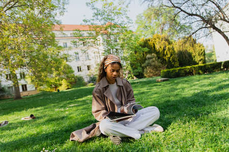 young african american woman in headscarf and stylish trench coat sitting on green grass and reading...
