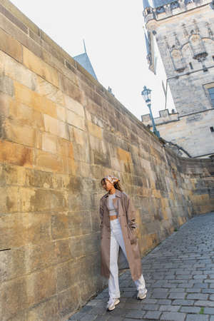 full length of stylish african american woman in trench coat walking near ancient wall in pragueの写真素材