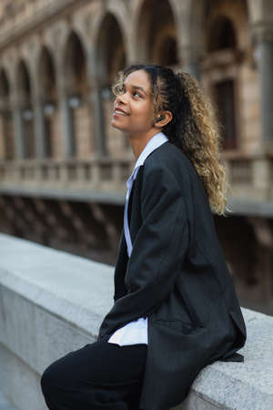 pleased african american woman in oversize suit listening music in wireless earphone on urban street...