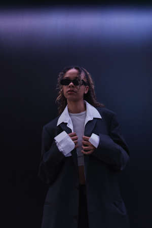 curly african american woman in sunglasses adjusting oversize blazer near dark blue wallの写真素材