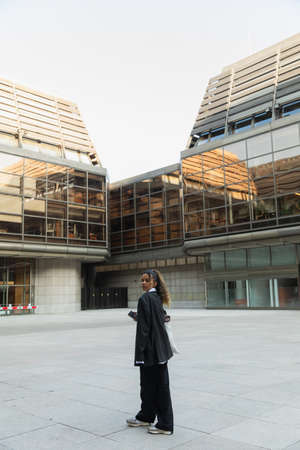 african american woman in oversize suit listening music in wireless earphone and walking on urban...