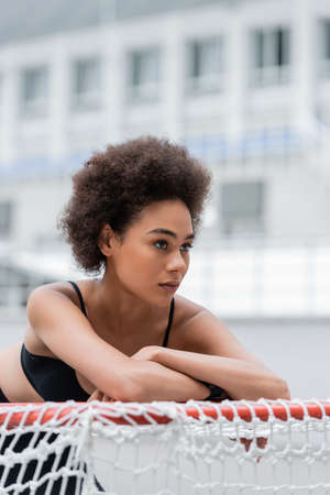 athletic african american woman with crossed arms leaning on sports gates and looking awayの写真素材