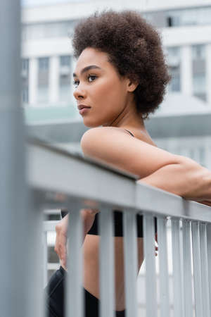sportive african american woman leaning on fence and looking away on blurred foregroundの写真素材