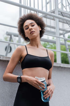 low angle view of sportive african american woman with sports bottle and fitness tracker looking at...