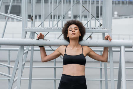 pretty and athletic african american woman in black sports bra looking at camera near fence