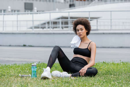 african american woman in black sportswear sitting near sports bottle and dumbbells on lawn