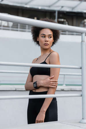 slender african american woman in black sportswear and fitness tracker looking away near fence