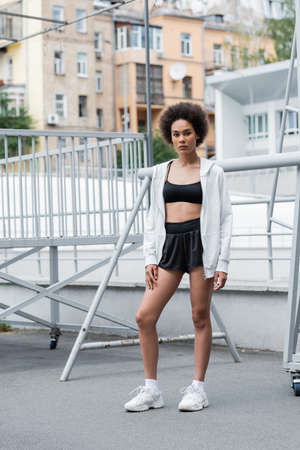 full length of african american woman in black sportswear and white hoodie standing on city stadiumの写真素材