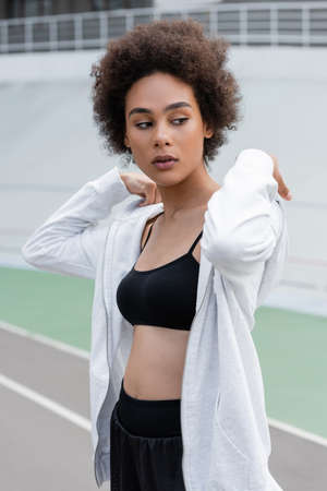 sportive african american woman in black sports bra and white sports jacket looking away