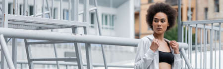 african american sportswoman in white hoodie and black sports bra near fence on stadium, bannerの写真素材