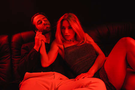 Passionate woman touching boyfriend on leather couch with red lighting isolated on blackの写真素材