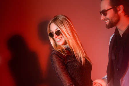 Smiling man in sunglasses looking at cheerful girlfriend during party on red background
