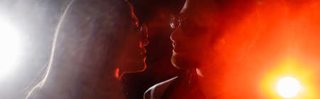 side view of couple in eyeglasses looking at each other on black background with bright light,...