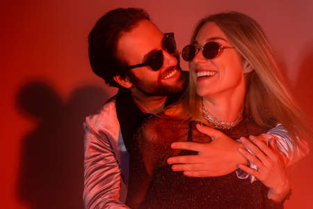 Young man in sunglasses embracing stylish girlfriend during party on red background