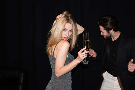 Sexy woman in dress holding champagne while dancing near boyfriend isolated on black