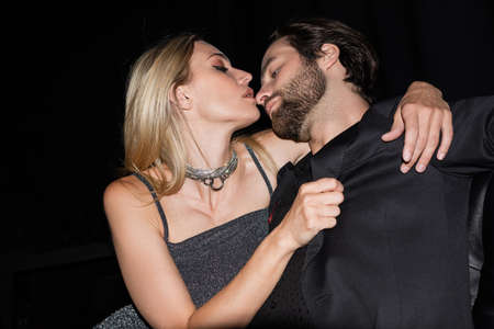 Seductive woman hugging bearded boyfriend in jacket on leather couch isolated on black