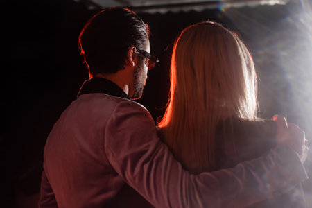 back view of man in trendy blazer and eyeglasses hugging blonde woman on black background with light