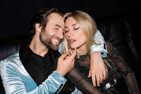 Cheerful man touching stylish girlfriend during party on leather couch isolated on blackの写真素材