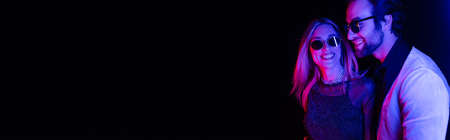 Smiling woman in sunglasses standing near stylish man isolated on black with colorful lighting, bannerの写真素材