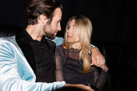 Stylish man hugging blonde girlfriend on leather couch during party isolated on black