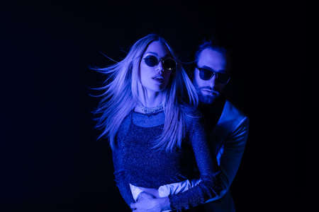 Man in sunglasses hugging blonde girlfriend during party isolated on black with blue lighting