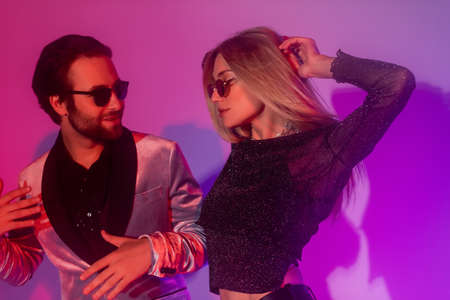 Stylish couple in sunglasses dancing during party on purple background