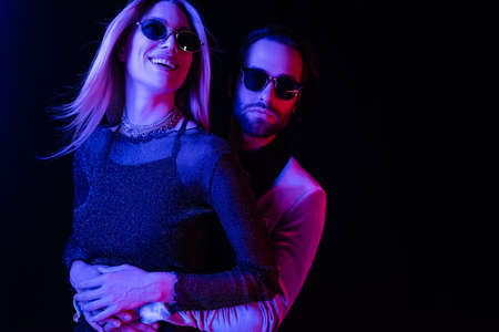 Young man in jacket hugging smiling girlfriend in sunglasses isolated on black with blue lighting