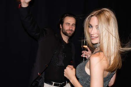 Smiling blonde woman holding glass of champagne while boyfriend dancing isolated on black
