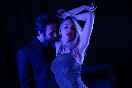Passionate man standing near sensual girlfriend in dress with blue lighting on black background