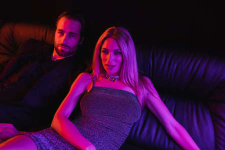 Seductive blonde woman looking at camera near boyfriend on leather couch with lighting isolated on...