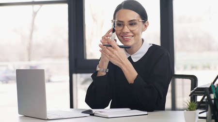 smiling businesswoman in eyeglasses holding pen and talking on mobile phone near laptop