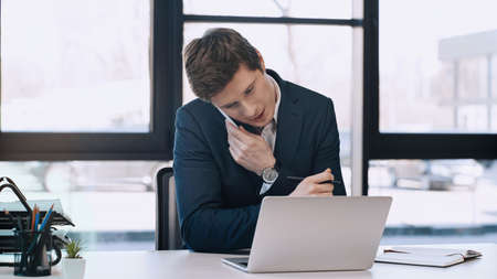 businessman holding pen and talking on mobile phone near laptopの写真素材