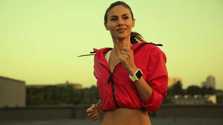 Smiling sportswoman with fitness tracker running on roof in evening