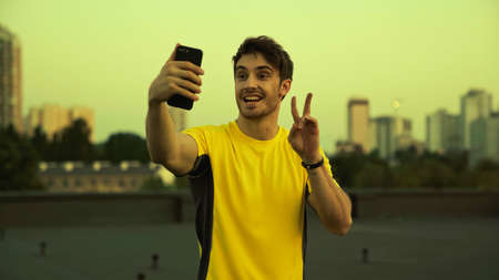 Smiling sportsman taking selfie on smartphone and gesturing on roof in eveningの写真素材