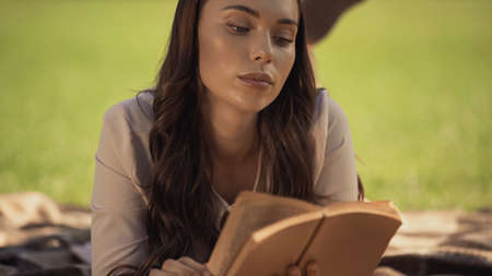 Brunette woman reading book while lying on blurred blanket in park