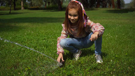 Red haired girl playing with automatic water sprayer in summer parkの写真素材