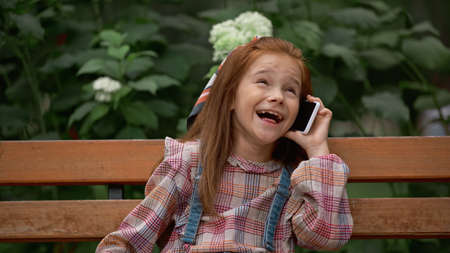 Positive child talking on smartphone while sitting on bench in park