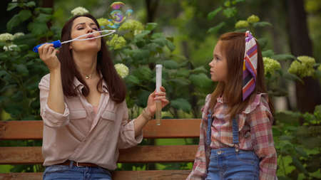 Woman blowing soap bubbles near redhead daughter on bench in parkの写真素材