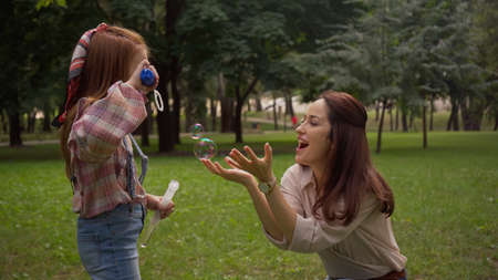 Positive woman standing near daughter blowing soap bubbles in park