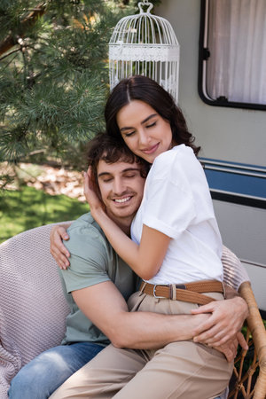 Brunette woman hugging boyfriend with closed eyes on armchair near camper vanの写真素材