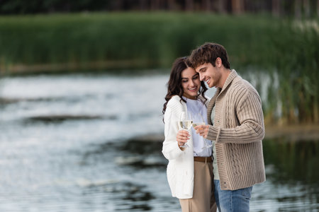Cheerful couple in knitted cardigans toasting wine near blurred riverの写真素材