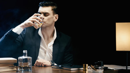 Businessman in suit drinking whiskey near books and ashtray on table isolated on blackの写真素材