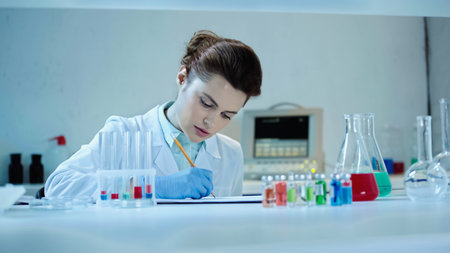 pretty scientist in white coat writing down result near test tubes and flasks with liquid in...