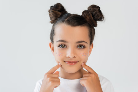 portrait of cheerful girl with lip gloss and sparkling glitter stars pointing at face isolated on greyの写真素材
