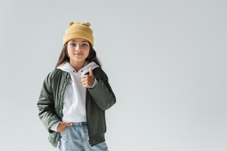 cute girl in beanie hat and stylish autumnal outfit posing with hand in pocket and showing thumb up isolated on greyの写真素材