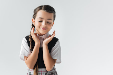 portrait of happy schoolgirl with closed eyes gesturing isolated on greyの写真素材