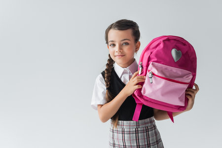 cute schoolgirl in uniform holding pink backpack and smiling isolated on greyの写真素材
