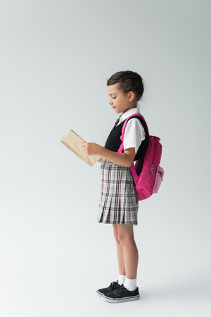 side view of smart schoolgirl in uniform reading book and standing and reading book on greyの写真素材