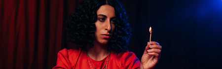 young fortune teller looking at burning palo santo stick on dark background with red drape, bannerの写真素材