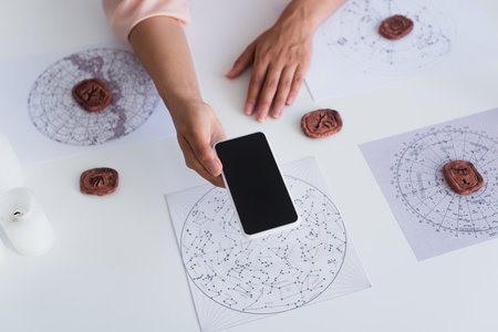 top view of cropped astrologist holding mobile phone with blank screen near celestial charts and clay runesの写真素材